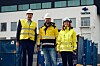Tre arbeidere i hjelmer og refleksjakker står foran containere ved en industriell bygning med NorSea-logo.