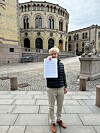 En mann står på en steinlagt plass foran en historisk bygning og holder opp et ark mot kamera.