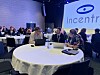 Mennesker sitter ved bord i en konferansesal med scenelys og en stor skjerm med Incentra-logo.