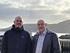 To menn står side om side utendørs på et fartøy med fjord og fjell i bakgrunnen.
